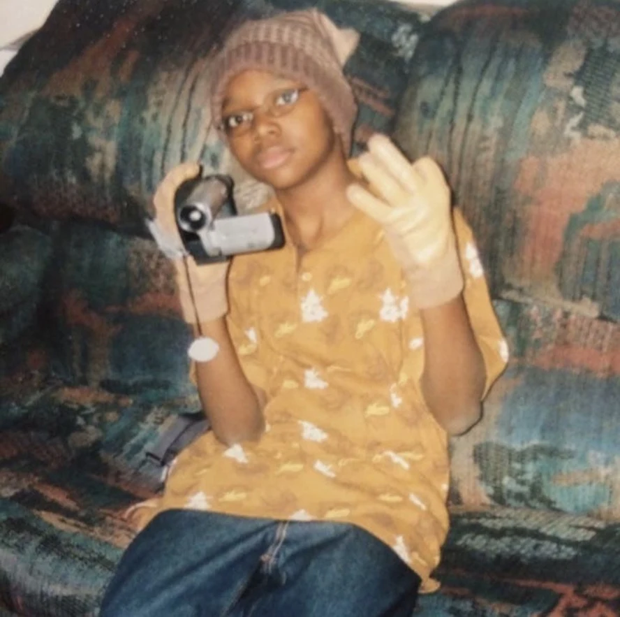 A young Rydrea Walker on a couch, holding a camcorder up to film